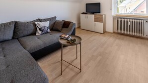 Uma sala de estar moderna com um sofá seccional, uma pequena mesa de centro, uma TV sobre um móvel e uma grande janela com vista para o telhado.