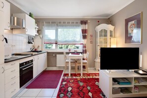 A compact kitchen with white cabinets, a stove, a dining table with chairs, a TV, and a window with curtains.
