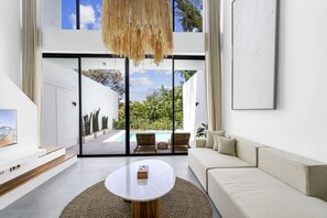 A modern living room with a large window, a white sofa, a coffee table, and a view of an outdoor pool area. - Villa Finity A by Hombali (Canggu)