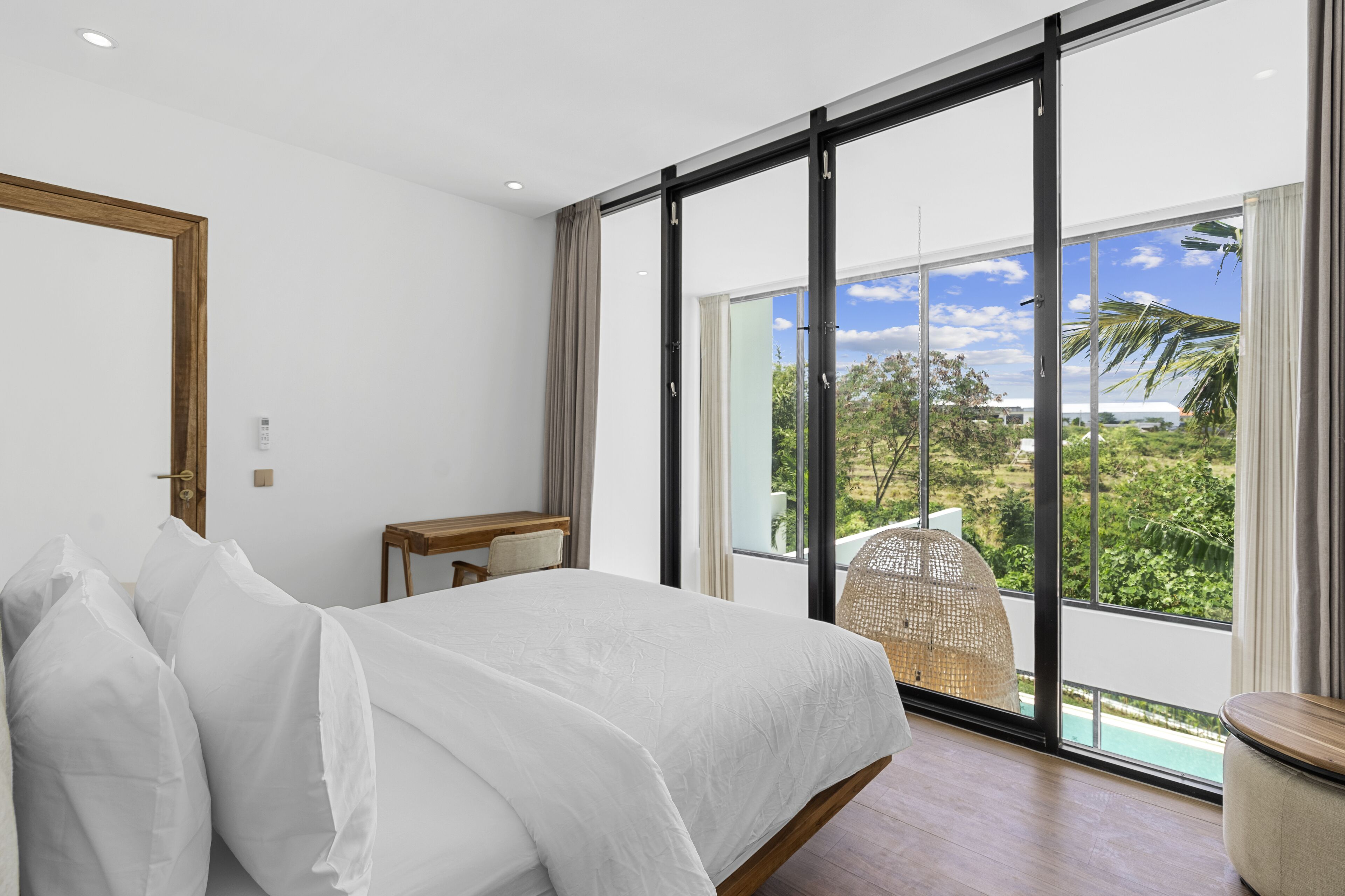 A modern hotel room with a large bed, a small desk, and a view of a tropical landscape through floor-to-ceiling windows.