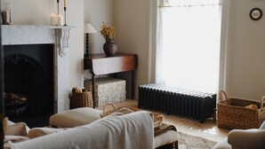 A cozy living room with a fireplace, a radiator, and a window with curtains. - The Nomad House, Ramsgate (Ramsgate)