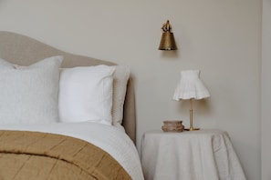 Una cama bien hecha con cabecera, una mesita de noche con lámpara, y una lámpara de pared.