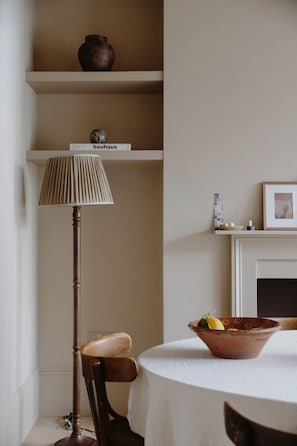 Una habitación con una mesa redonda, una lámpara, una silla, una estantería con un jarrón y un cuadro enmarcado.