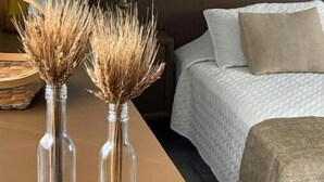 Et soveværelse med en seng, et natbord, en lampe og to klare glasvaser med tørret hvede på et træbord.