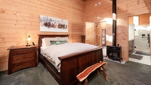 A wooden cabin bedroom with a bed, bedside table, lamp, and a painting of horses. - Modern Mountain Retreat w/ Amazing Views • Hot Tub • Deck • Pets Welcome (Idyllwild-Pine Cove)