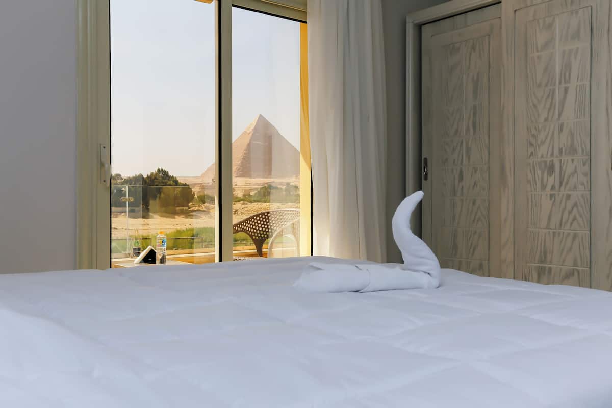A hotel room with a bed, a view of the pyramids, and a wooden wardrobe.