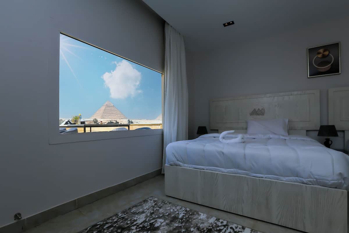 A bedroom with a large window, a bed, a nightstand, and a view of a pyramid.