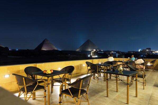 Un espace de restauration en plein air avec des tables et des chaises, une vue sur les pyramides en arrière-plan et un paysage urbain illuminé la nuit.