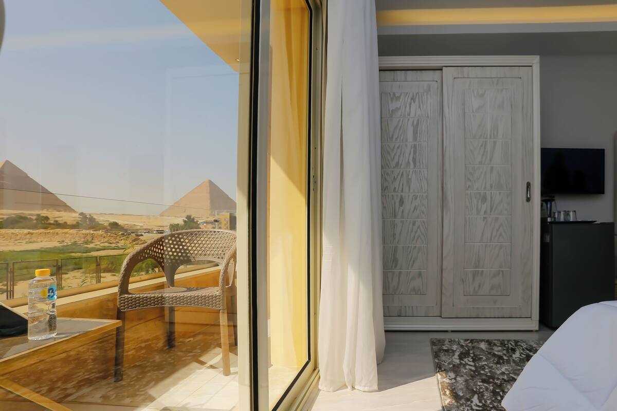 A hotel room with a view of the pyramids, a bed, a TV, and a dining area with a chair and table.
