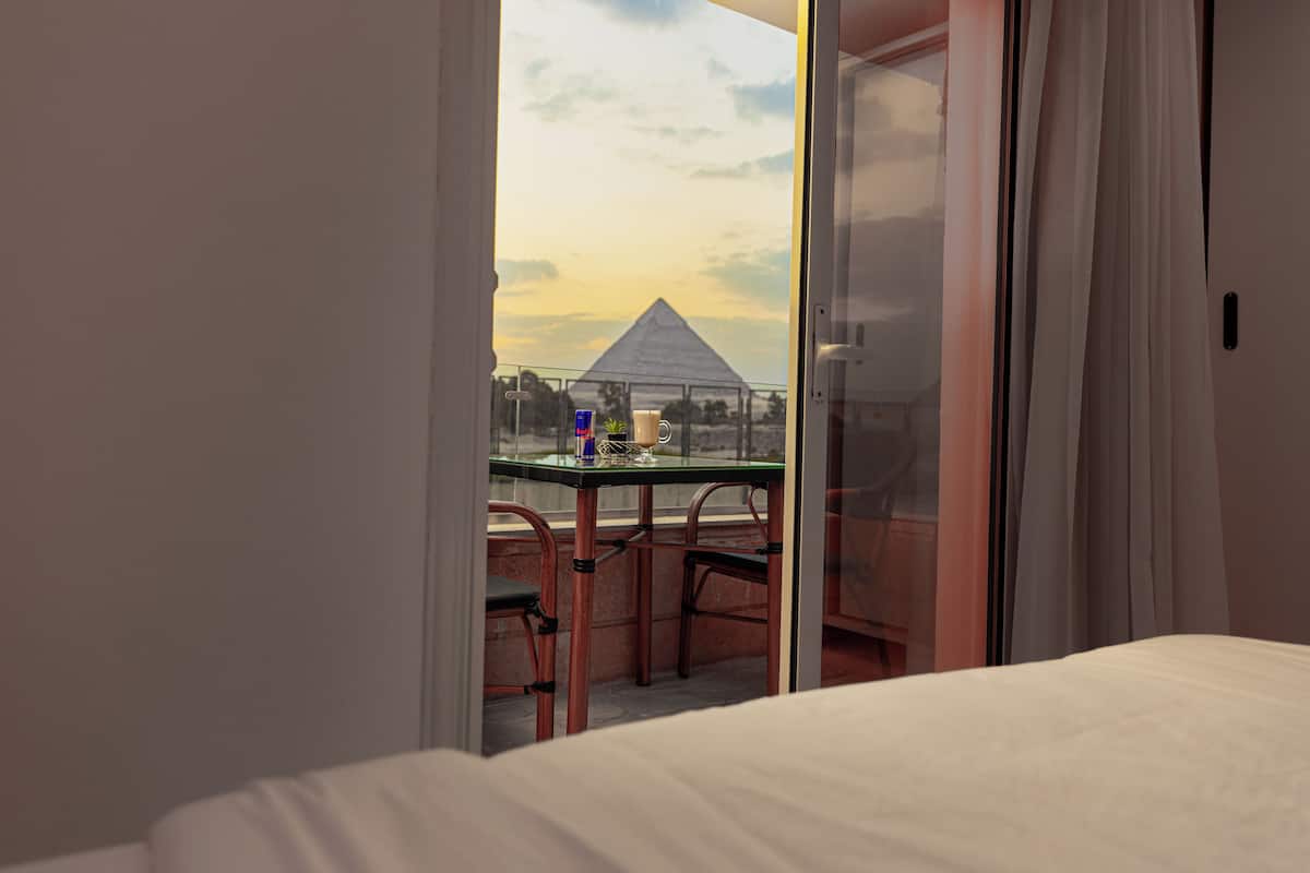 A room with a bed, a glass table with chairs, and a view of a pyramid through a glass door.