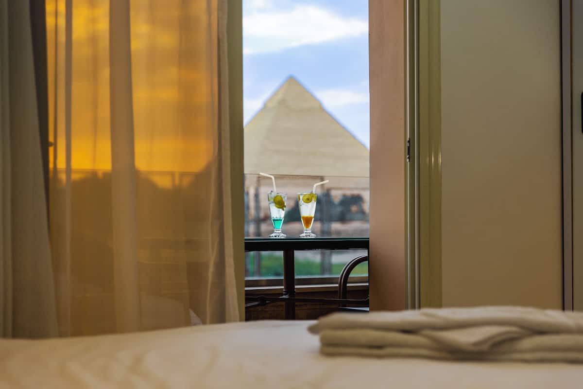 A hotel room with a bed, a window with curtains, and a view of a pyramid.