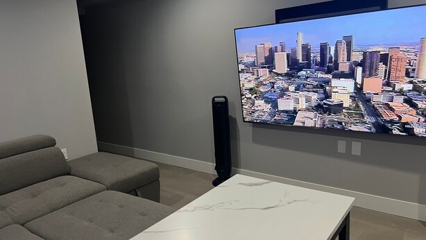 Una sala moderna con un televisor de pantalla plana grande mostrando un paisaje urbano, un sofá gris y una mesa de centro de mármol.