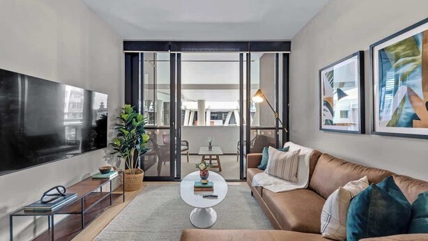 A modern living room with a flat-screen TV, a sofa, a coffee table, and a dining area visible through glass doors.