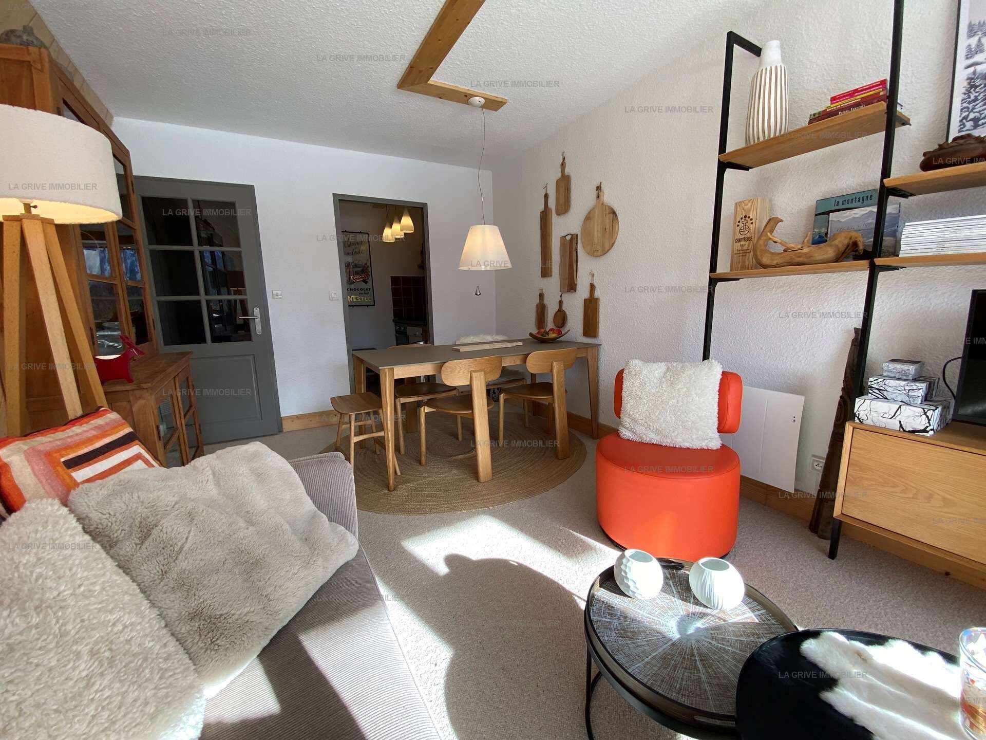 Ein modernes Wohnzimmer mit einer Couch, einem Esstisch mit Stühlen, einem roten Hocker und einem Bücherregal.