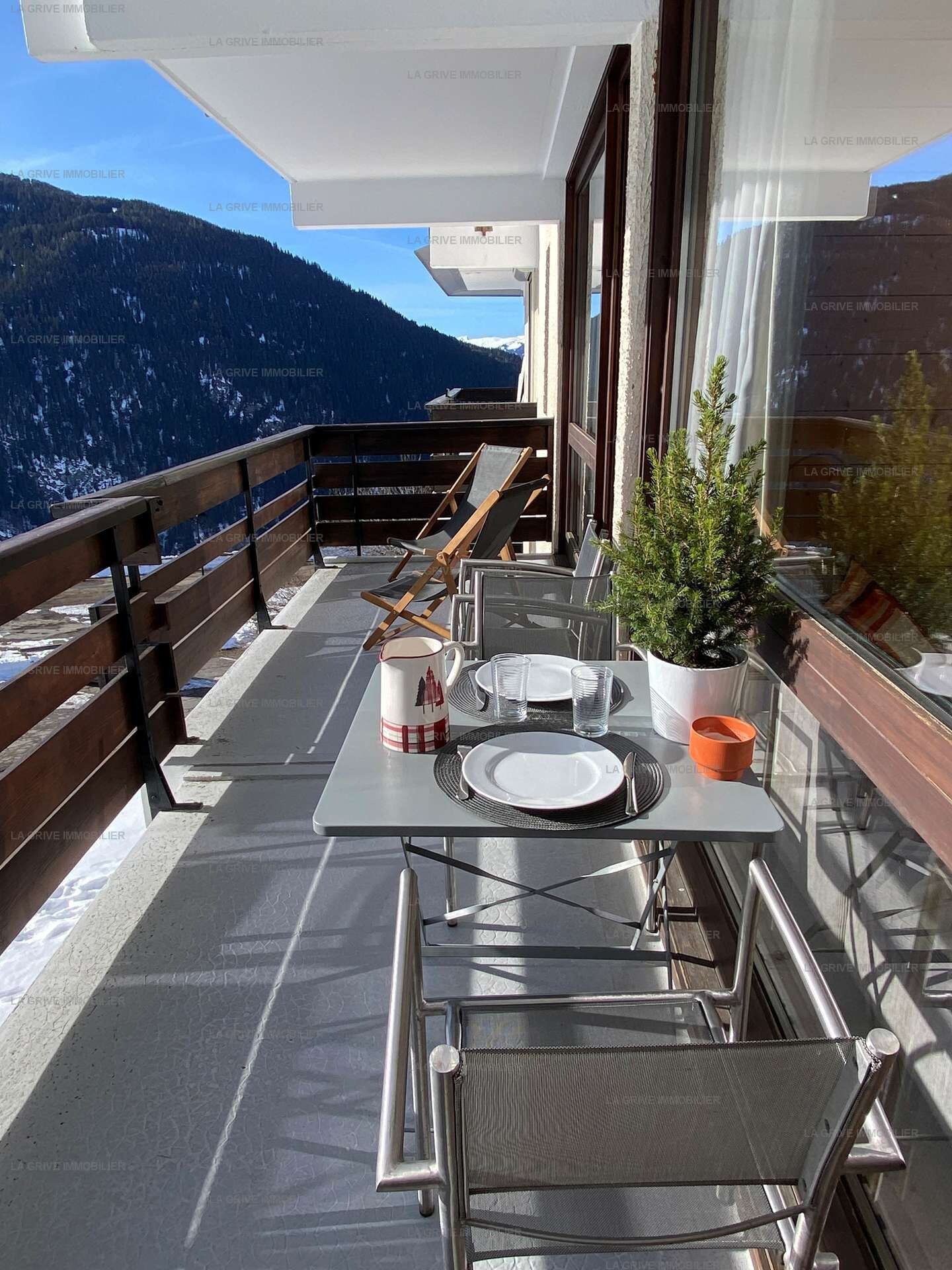 Varanda com mesa posta para dois, um vaso de planta e vista para uma paisagem montanhosa.