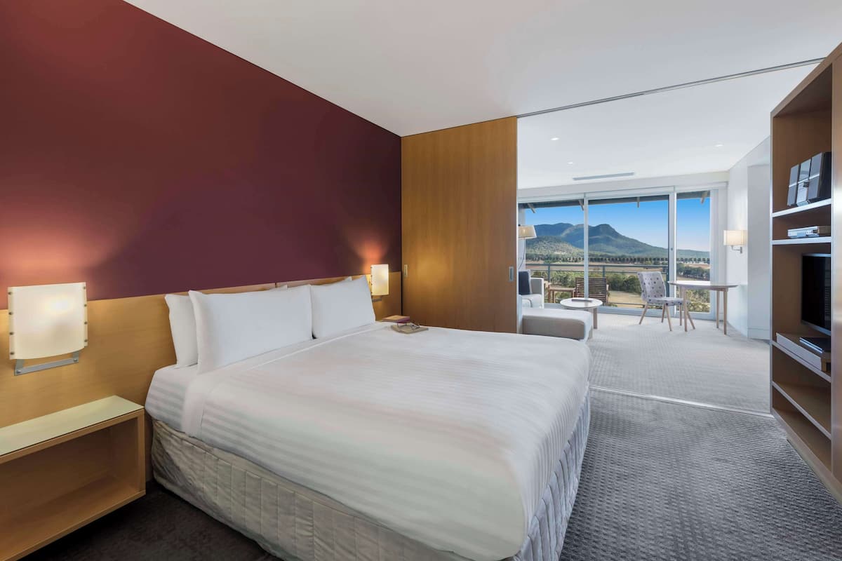 A hotel room with a large bed, a TV, a desk, and a balcony with a mountain view.