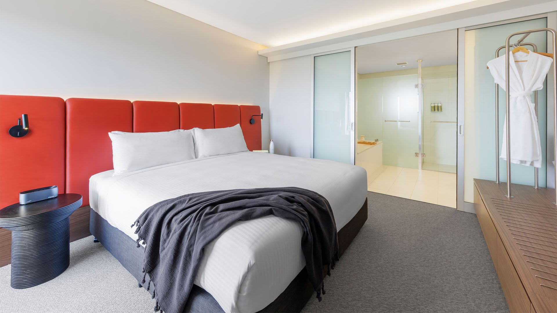 A modern hotel room with a large bed, a red accent wall, a bedside table with a lamp, and a glass-enclosed bathroom.