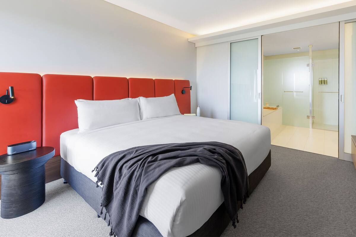 A modern hotel room with a large bed, a red accent wall, a bedside table with a lamp, and a glass-enclosed bathroom.