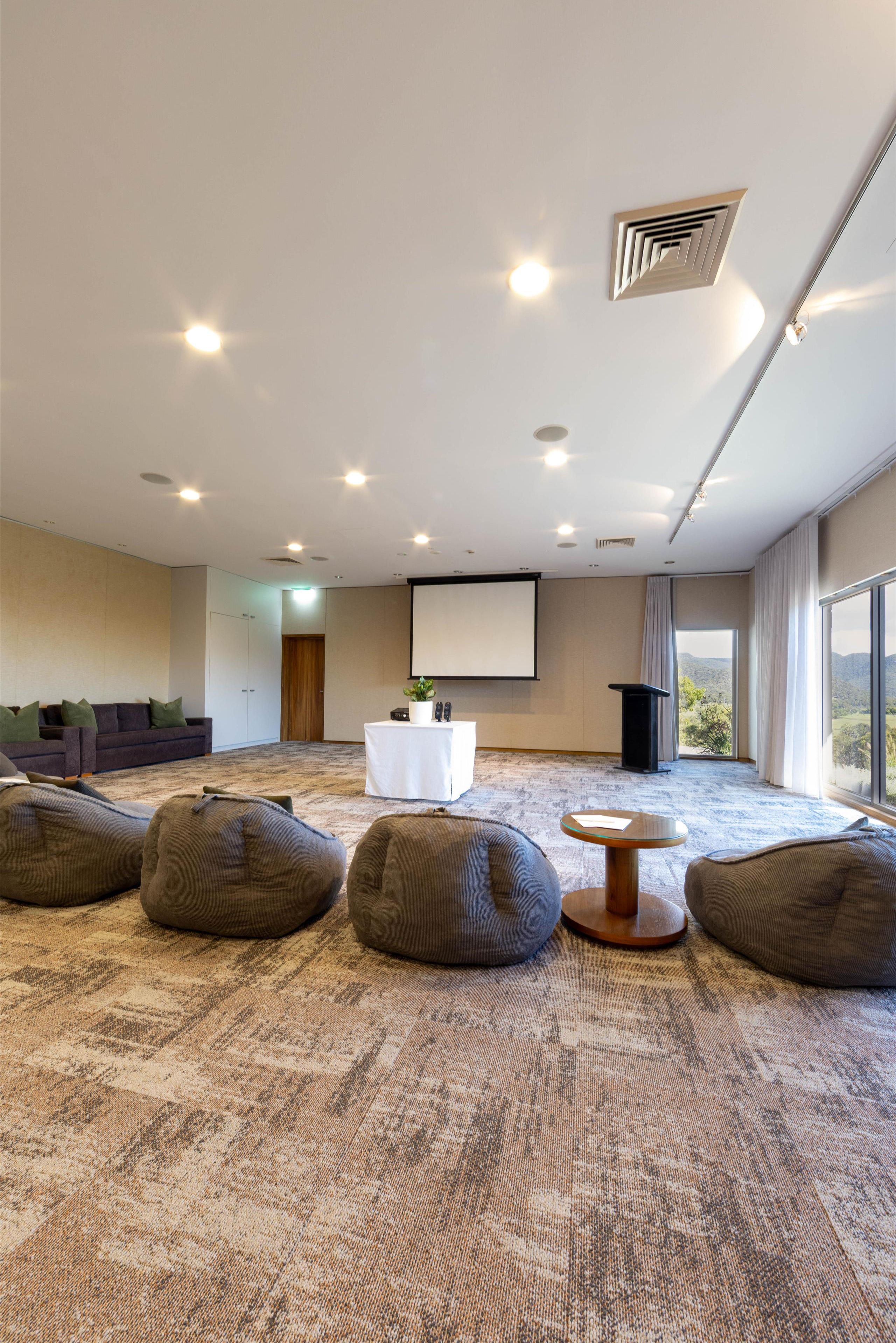 A spacious room with a large screen, bean bag seating, a small table, and a view through a large window.
