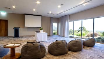 Un salón de conferencias con suelo alfombrado, pufs, una pantalla de proyector, un atril y una mesa con una planta.