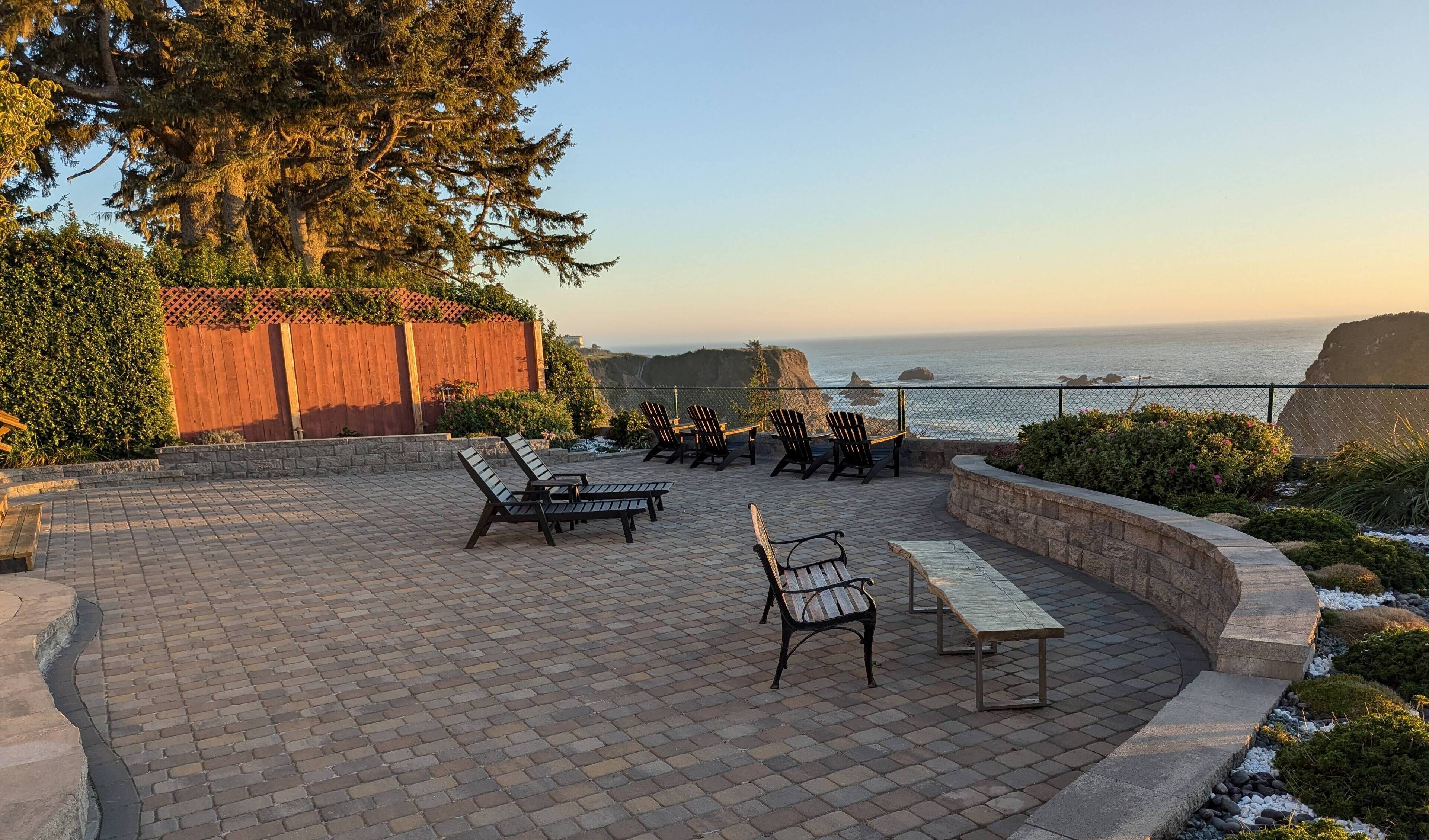 An outdoor patio with brick flooring, wooden chairs, and a bench, overlooking a coastal view with a wooden fence and greenery.