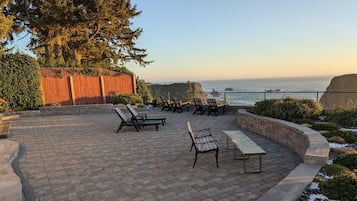 An outdoor patio with brick flooring, wooden chairs, and a bench, overlooking a coastal view with a wooden fence and greenery.