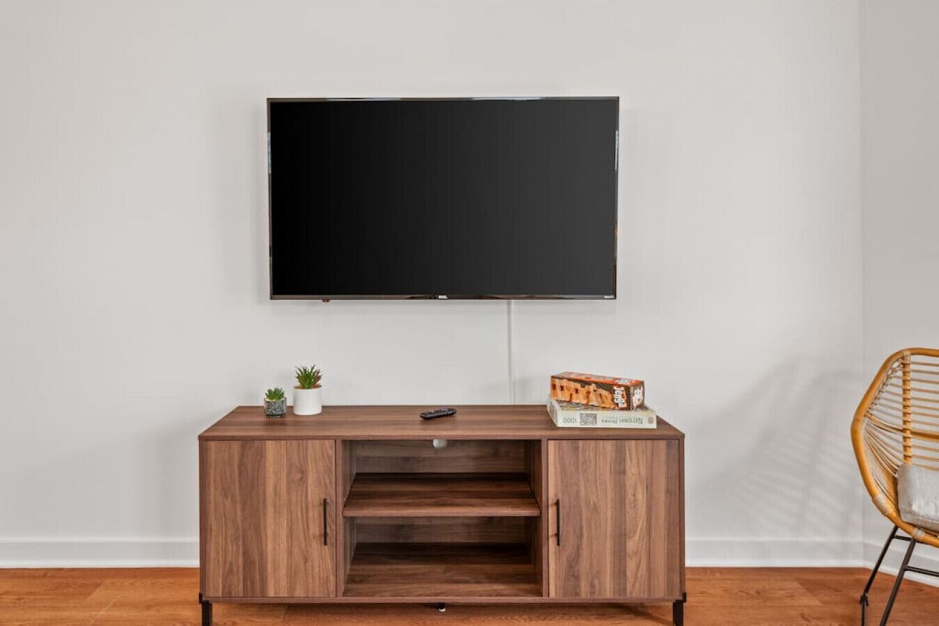 A modern living room with a flat-screen TV mounted on the wall, a wooden entertainment center with a remote control and a game box, a small potted plant, and a wicker chair.