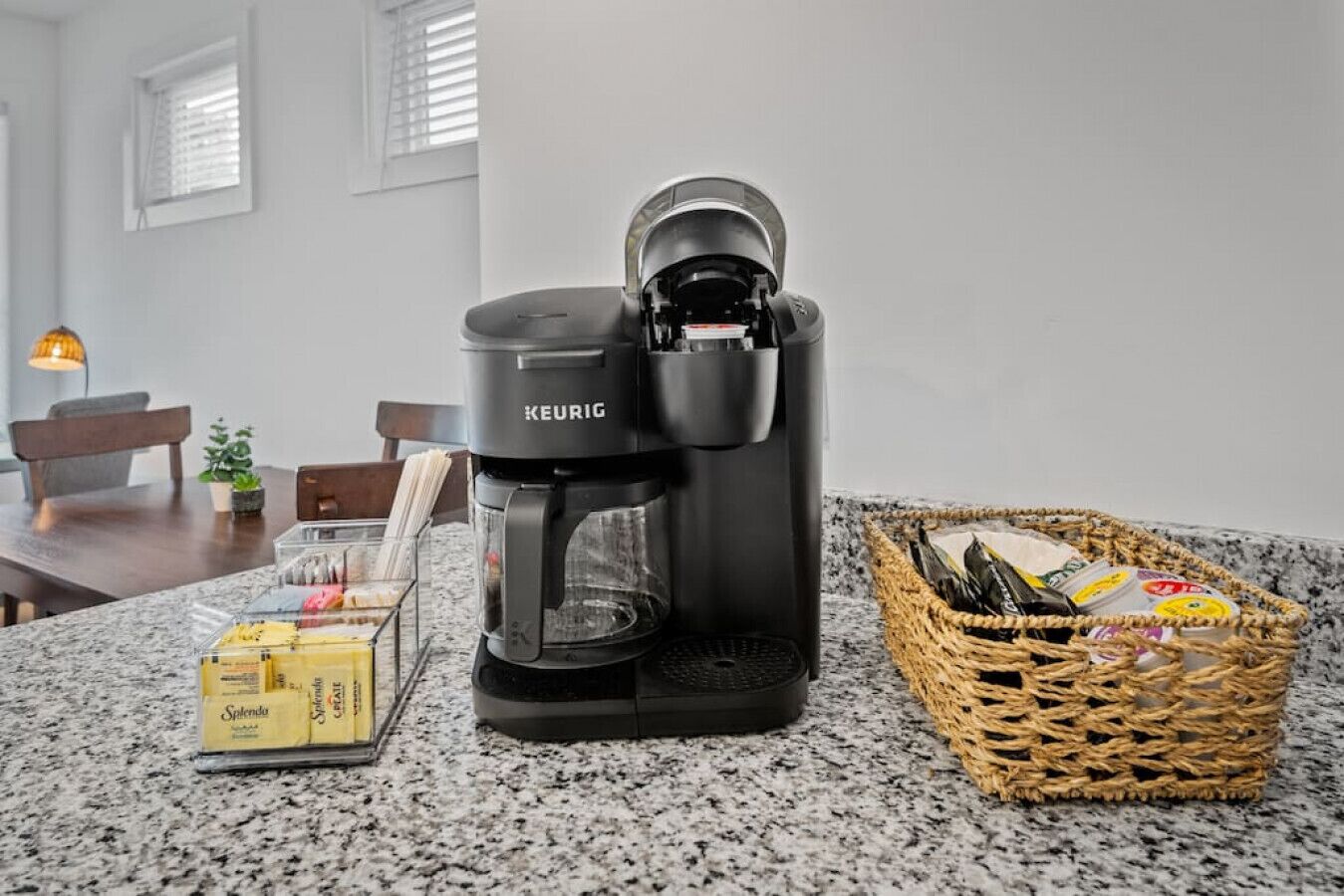 A Keurig coffee maker on a kitchen counter with a basket of coffee pods and a box of Splenda sugar.