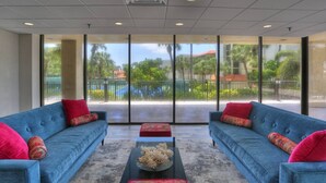 A spacious room with two blue sofas, a black coffee table, and a large window offering a view of a courtyard with palm trees and a pool. - Gulf Views From Every Room! Heated Pool. Direct Beach Access.  Recreation Room. Beautiful Beach. (Treasure Island)