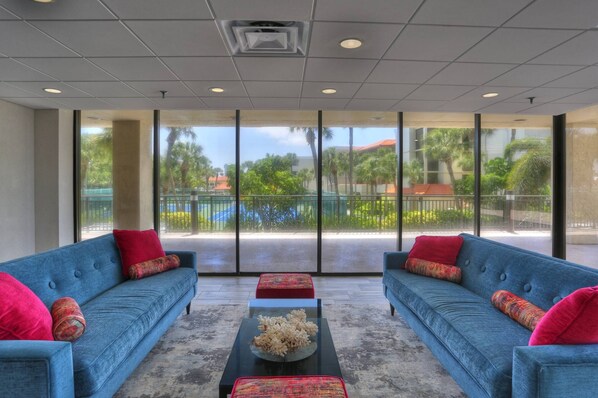 A spacious room with two blue sofas, a black coffee table, and a large window offering a view of a courtyard with palm trees and a pool.