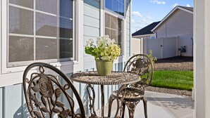 A patio with a round metal table and chairs, a potted plant with white flowers, and a view of a white house with a screened window.