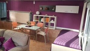 Habitació amb una paret morada, un llit blanc, un sofà beix amb coixinets morados i una taula de vidre, juntament amb cadires taronja.