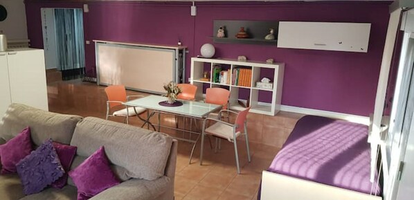 A room with a purple wall, a white bed, a beige sofa with purple cushions, a glass table, and orange chairs.