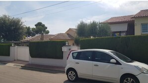 Un cotxe blanc estacionat al carrer, amb una entrada tancada per una reixa i un edifici amb teulada enrajolada al fons.