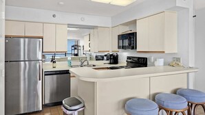 A modern kitchen with a stainless steel refrigerator, a dishwasher, a microwave, and a coffee maker. There are three bar stools and a trash bin. - San Luis Beauty in The Beautiful San Luis Resort Pet Friendly (Galveston)