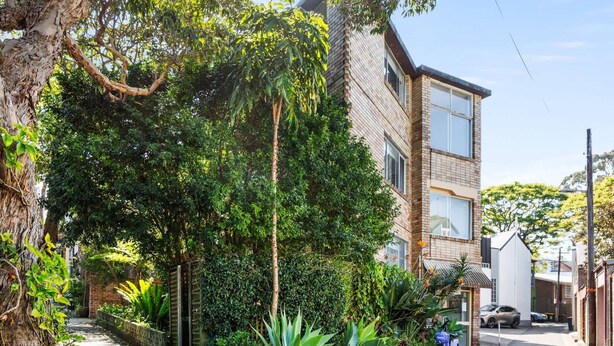 Un edificio residencial con fachada de ladrillo, ventanas grandes y un jardín con diversas plantas y árboles.