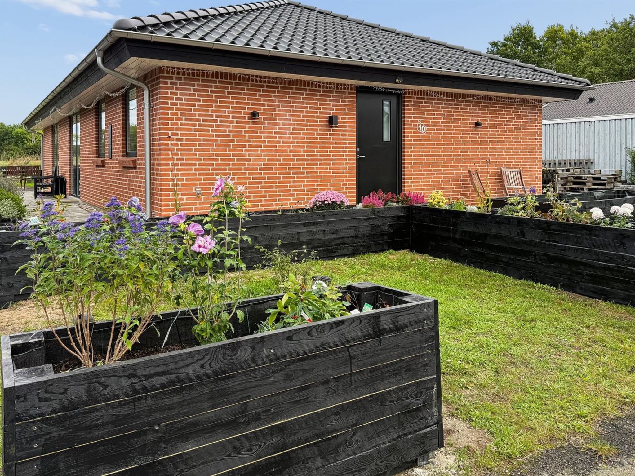 A brick building with a black door, surrounded by raised garden beds with various plants.