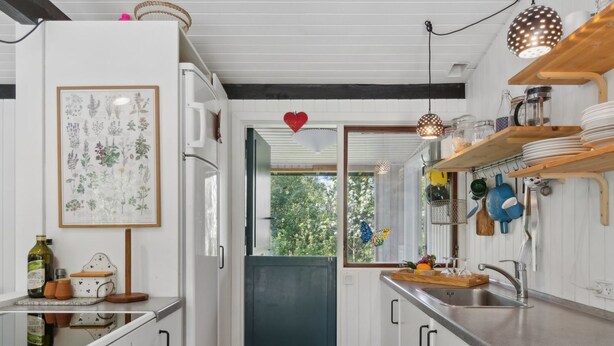 Une cuisine compacte avec des meubles blancs, un plan de travail en bois et une vue sur un espace extérieur verdoyant, accessible par une porte ouverte.