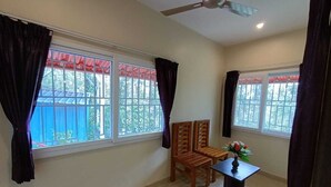 Una habitación con dos ventanas, un ventilador de techo, una mesita con un florero y dos sillas de madera.