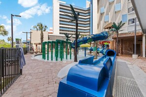 A playground with slides and climbing structures, surrounded by buildings and palm trees.