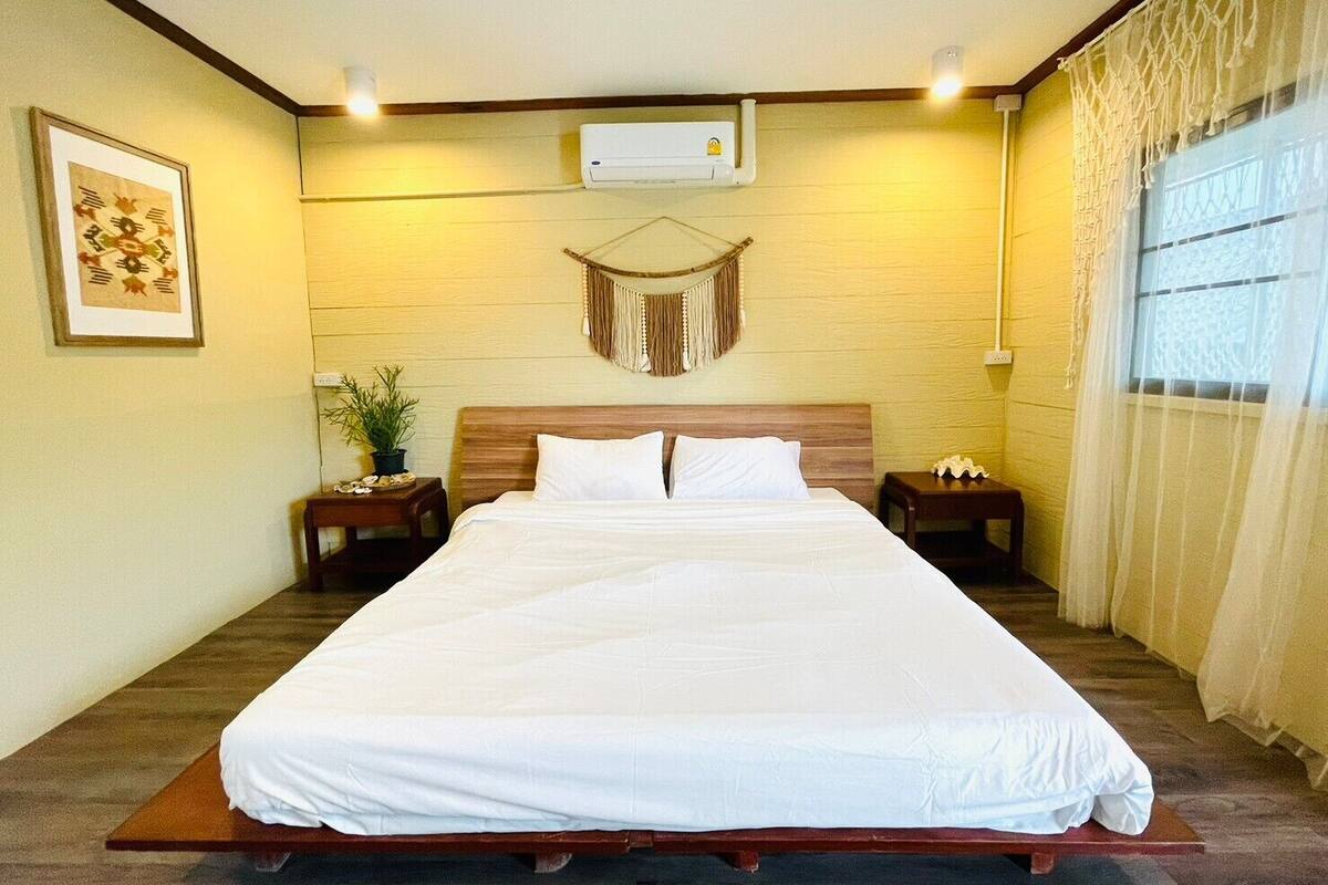 Uma cama grande com roupa de cama branca, cabeceira de madeira, duas mesas de cabeceira, um ar condicionado fixado na parede, um pendente de macramé e um quadro na parede à esquerda.
