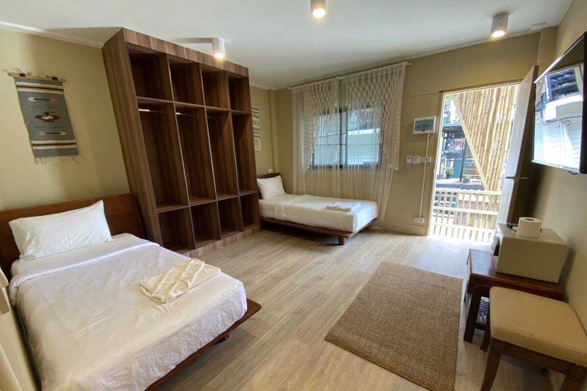 Quarto de hotel com duas camas, um guarda-roupa de madeira, uma secretária com cadeira, uma janela com cortinas e vista para o exterior.