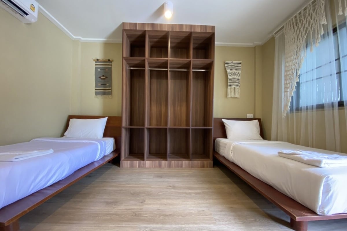 Habitació d'hotel amb dos llits individuals, un armari de fusta, un escriptori petit i una finestra amb cortines primes.