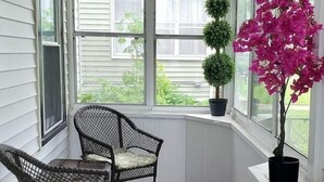 Eine überdachte Veranda mit zwei Korbstühlen, einem Blumentopf und einem großen Fenster mit Blick auf einen Garten.