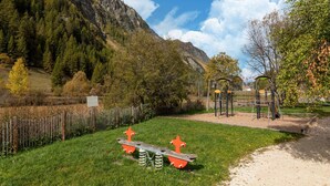 Un terrain de jeux avec des balançoires et une structure d’escalade, entouré d’une clôture en bois et avec une chaîne de montagnes en arrière-plan.