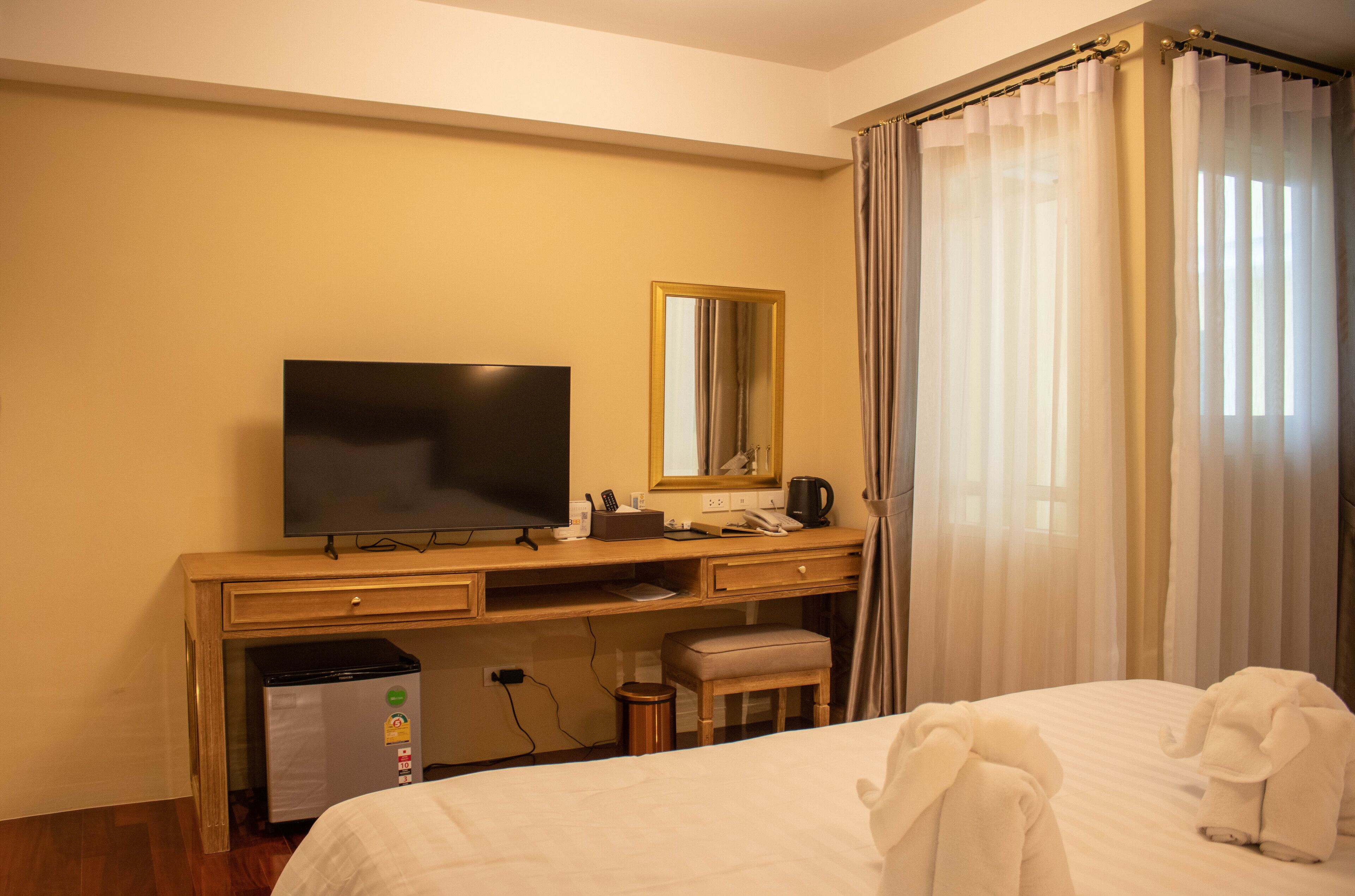 Habitación de hotel con cama, escritorio con televisor, espejo, taburete pequeño y ventana con cortinas.