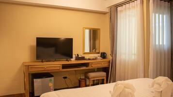 Ett hotellrum med en säng, ett skrivbord med en tv, en spegel, en liten pall och ett fönster med gardiner.