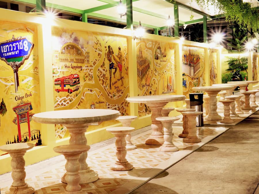Una hilera de mesas blancas con patas ornamentadas, frente a una pared con murales amarillos que muestran varios puntos de referencia y un autobús rojo.