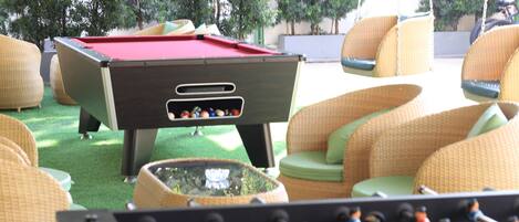 A pool table with a red felt surface, surrounded by wicker chairs with green cushions.