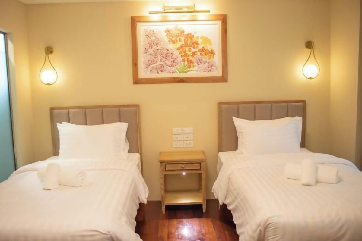 A hotel room with two beds, a small bedside table, a framed floral artwork on the wall, and wall-mounted light fixtures.